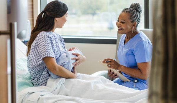 A mother in maternity room cradles her newborn as an older nurse gives support